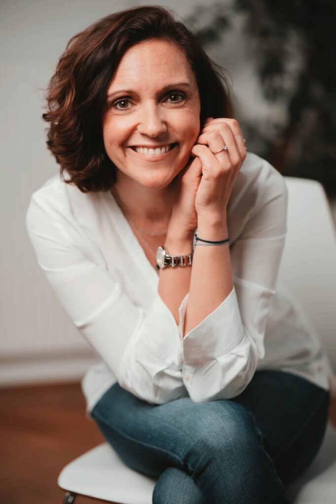 Olivia Prader sitzt entspannt mit verschränkten Armen und offenem Lächeln – Symbol für persönliche 1:1-Trainings in der Geldsprechstunde.